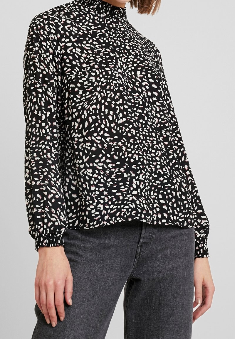 Femme portant un chemisier noir à manches longues avec des taches abstraites blanches et roses, associé à un jean gris foncé, sur un fond clair uni.