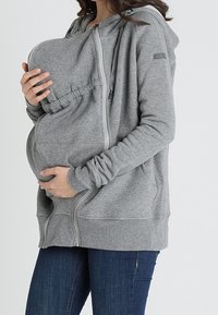 Donna che indossa una felpa grigia con zip e tasca integrata, tiene un bambino piccolo vicino al petto.