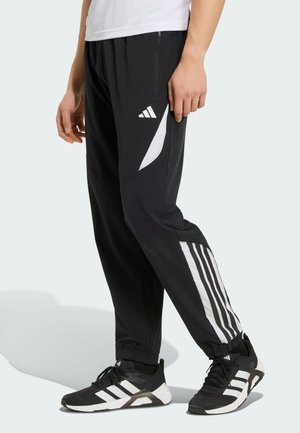 Person trägt schwarze Adidas Trainingshose mit weißen Streifen und schwarze Adidas Sneakers mit weißen Streifen, stehend auf hellem Hintergrund.