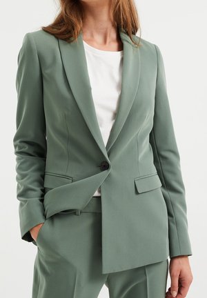 Frau trägt ein hellgrünes, maßgeschneidertes Sakko mit einem Knopf, der über einem weißen Oberteil geschlossen ist, dazu passende Hosen und hat die Hände in den Taschen.