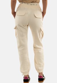 Pantalones cargo beige con cintura y puños elásticos, que presentan dos bolsillos laterales y una textura suave y lisa. Los acentos incluyen un logo en la cinturilla.