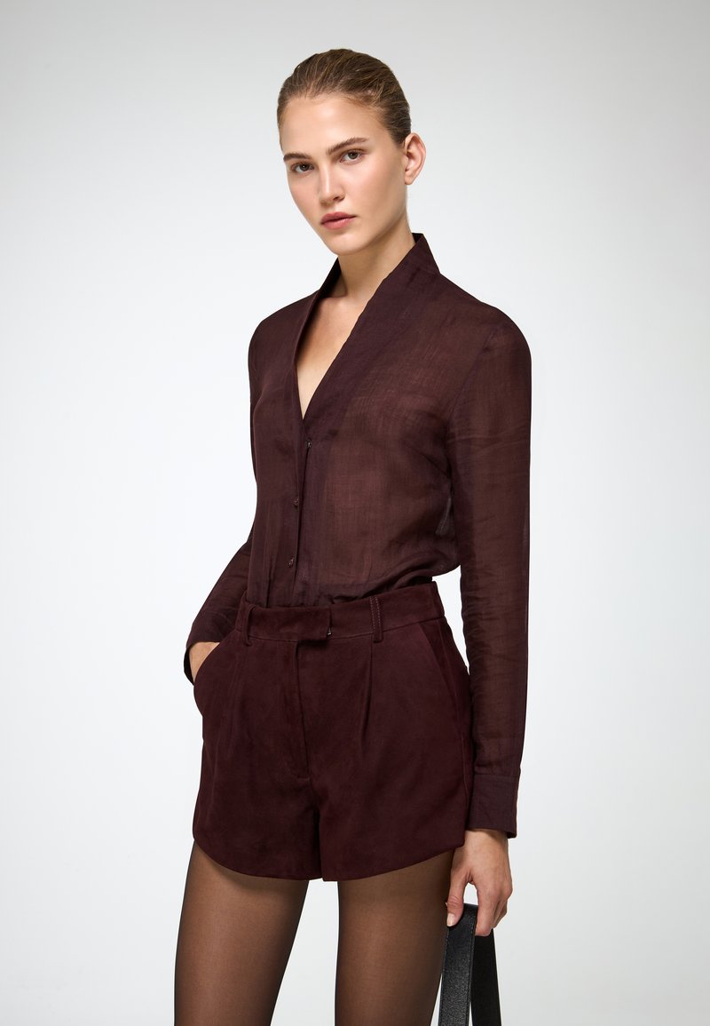 Camicia trasparente bordeaux con colletto abbinata a pantaloni corti in suede a vita alta dello stesso colore. Una sottile cintura nera mette in risalto il punto vita.