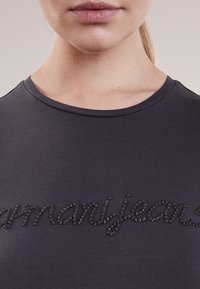 T-shirt grigio scuro con una texture liscia e scollatura a girocollo, decorato con strass neri che formano il testo "manyjeans."