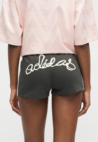 Άτομο με σκούρα σορτς που έχει λευκό λογότυπο σε καλλιγραφική γραφή στο πίσω μέρος και ένα ανοιχτό ροζ πουκάμισο με διακριτικό γεωμετρικό μοτίβο, από πίσω.