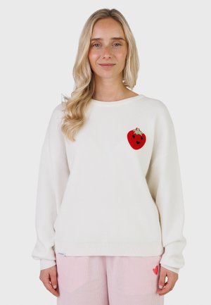 Giovane donna con lunghi capelli biondi che indossa un maglione bianco con una toppa a forma di fragola rossa e pantaloni rosa chiaro con un piccolo cuore rosso.