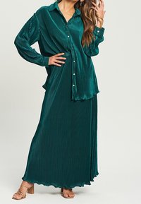 Abito maxi plissettato color turchese con parte superiore in camicia a bottoni, maniche lunghe e gonna fluente. Tessuto morbido e dettaglio del bordo ondulato.