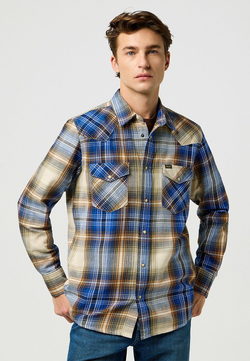 Camisa de manga larga a cuadros en azul, marrón y crema, con dos bolsillos en el pecho, yugo con puntas y diseño abotonado. Acabado de tela texturizada.