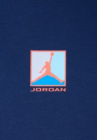 Námořnické tričko s zjednodušeným logem postavy ve skoku, orámované v korálové barvě, nad textem "JORDAN" ve shodné korálové barvě.