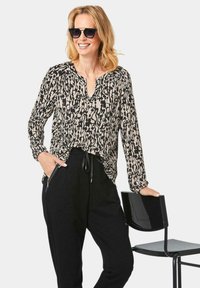 Zwart en beige gebloemde blouse met lange mouwen en een V-hals, gedragen met zwarte joggers. Model poseert naast een zwarte stoel, met zonnebril op.