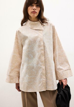 Femme portant une veste oversize beige à motifs floraux avec des manches larges, tenant un sac noir sur un fond uni.