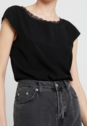 Vrouw draagt een zwarte mouwloze top met kanten randje bij de hals, ingestopt in een hooggesneden, uitgebleekte zwarte jeans.