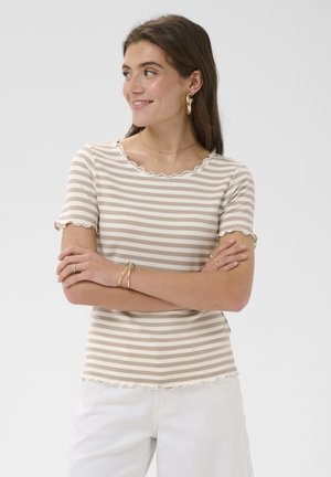 Naine, kellel on pikad pruunid juuksed, kannab beežide ja valgete triipudega lühikeste varrukatega särke, valgeid pükse, kuldseid rõngakujulisi kõrvarõngaid ja kuldset käevõru, naeratab käed rüpes.