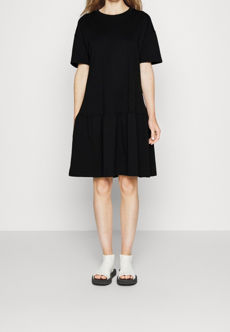Femme portant une robe noire ample à manches courtes avec une jupe froncée et des bottines blanches à bouts ouverts, debout devant un fond uni.