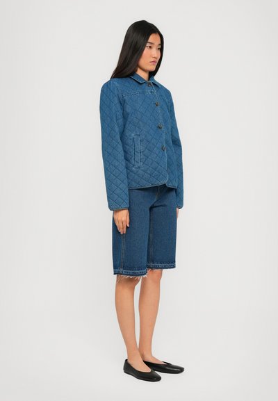 Femeie îmbrăcată într-o jachetă din denim albastru matlasat, pantaloni scurți din denim până la genunchi cu margini brut și pantofi negri cu talpă joasă, în fața unui fundal alb simplu.