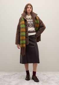 Manteau en peau retournée marron sur un pull crème à motifs, associé à une longue écharpe dégradée verte et jaune. Jupe en cuir noire et chaussures foncées.
