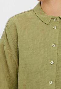 Light green button-up shirt with a textured fabric, collar, and four buttons visible down the front, showcasing a relaxed, oversized fit.