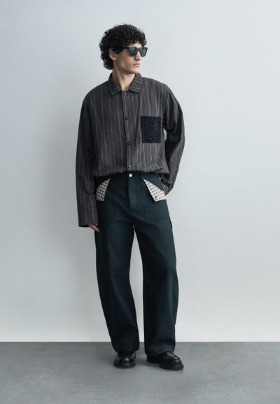Άνδρας που φοράει σκούρο πουκάμισο με λεπτές ρίγες και μαύρη υφή στην τσέπη, φαρδιά μαύρη παντελόνια με καρό ύφασμα να προεξέχει από τις τσέπες, μαύρα παπούτσια και γυαλιά ηλίου.