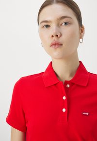 Mujer joven con piel clara y ojos azules que lleva pendientes de aro plateados y una camisa polo roja de Levi's con tres botones, fondo neutro.