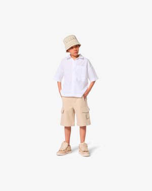 Chemise blanche à manches courtes, short cargo beige avec poches, et baskets beiges avec des accents texturés. Le mannequin porte un chapeau bob à motifs.