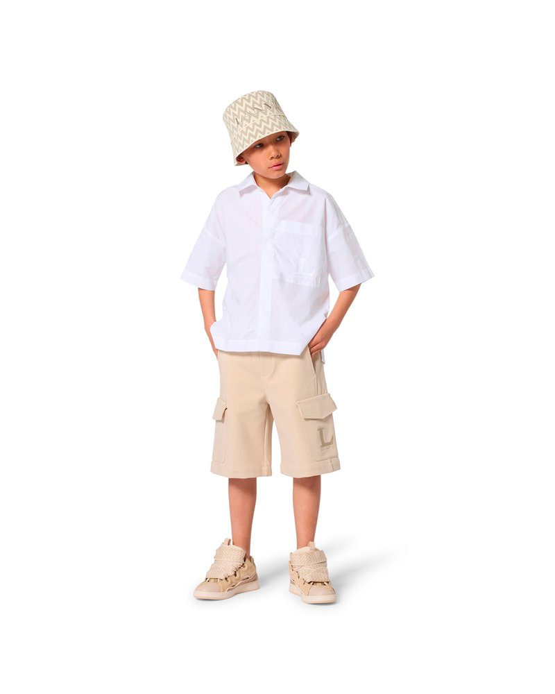 Chemise blanche à manches courtes, short cargo beige avec poches, et baskets beiges avec des accents texturés. Le mannequin porte un chapeau bob à motifs.