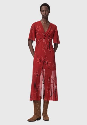 Frau steht in einem roten Midikleid mit Blumenmuster, flatternden Ärmeln, V-Ausschnitt, vorderem Schlitz und braunen kniehohen Wildlederstiefeln.