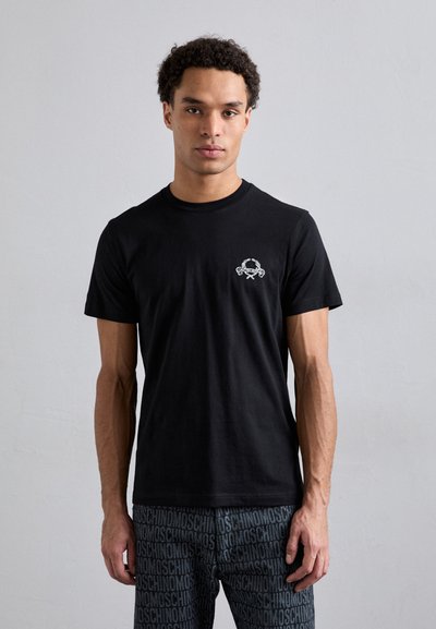 Schwarzes Baumwoll-T-Shirt mit Rundhalsausschnitt, mit einem weißen Logodetail auf der linken Brust. Das Modell trägt gemusterte graue Hosen.