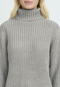 Grijs gebreid vest met een hoge geribbelde kraag, voorzien van een textuurpatroon en lange mouwen. Zacht, flexibel materiaal, ideaal om te combineren.