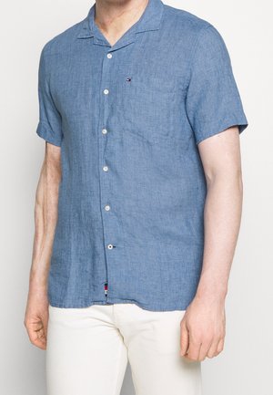 Muž stojící před neutrálním pozadím, oblečený v modré košili s krátkým rukávem na knoflíky s kapsou na hrudi a bílých kalhotách.