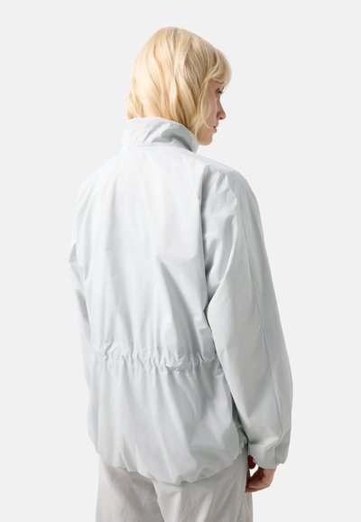 Blond persoon draagt een lichtgrijze, loszittende jas met een hoge kraag en een elastische tailleband, gezien van achteren tegen een effen achtergrond.