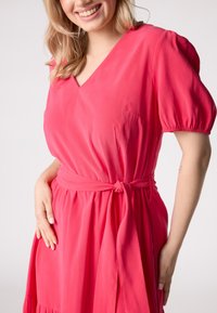 Robe rose avec un décolleté en V, des manches courtes bouffantes, une ceinture nouée à la taille et une jupe fluide avec une texture froncée.