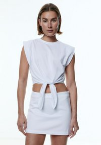 Witte cropped top met korte mouwen en een knoop aan de voorkant, gecombineerd met een witte minirok. Glad katoenen materiaal, strakke lijnen, minimalistisch ontwerp.