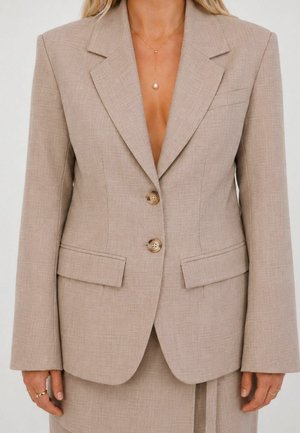 Beige skræddersyet blazer med to knapper, klaplommer og matchende nederdel, båret med en guldkæde med en perlevedhæng.