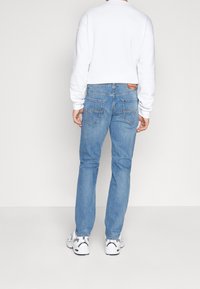 Person som bär ljusblå jeans, vita sneakers och en vit långärmad tröja, stående vänd bort mot en enfärgad ljus bakgrund.