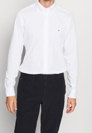 Mann trägt ein weißes langärmeliges Hemd und schwarze Hose, steht vor einem schlichten hellen Hintergrund.