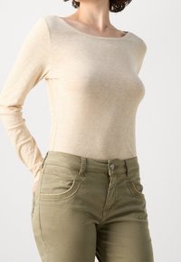 Beige top met lange mouwen en een brede halslijn, gecombineerd met olijfgroene, nauwsluitende spijkerbroeken met gestikte zakdetails en een ritssluiting aan de voorkant.