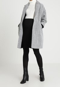 Grijs-wit jasje met een houndstooth-patroon en een oversized pasvorm, met een open voorkant, zijzakken, en gedragen over een zwarte outfit en laarzen.