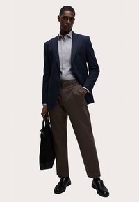 Blazer marine porté sur une chemise gris clair, associé à un pantalon marron ajusté et des chaussures noires. Tient un sac cabas noir. Texture de tissu lisse.