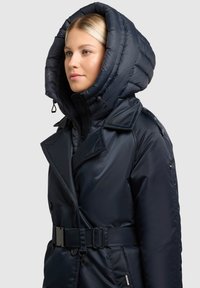 Temno modra puhovka z veliko strukturirano kapuco, zapiranjem na gumbe in pasom. Ima gladko površino in stranske žepe.