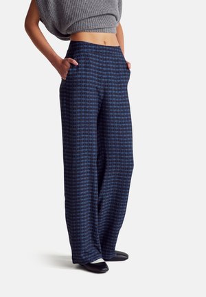 Femme portant un pantalon taille haute large bleu marine avec des motifs circulaires et un top gris court en maille, mains dans les poches, debout sur fond blanc.