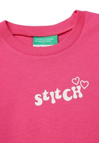 Růžové bavlněné tričko s bílým textem "stitch" a grafikou srdce, kulatým výstřihem a značkovou visačkou v zelené barvě uvnitř límce.