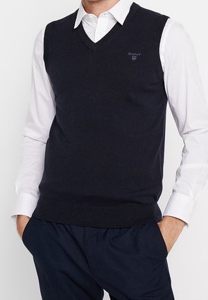 Homme portant une chemise blanche à col sous un pull sans manches bleu marine à col en V et un pantalon bleu marine, les mains dans les poches.