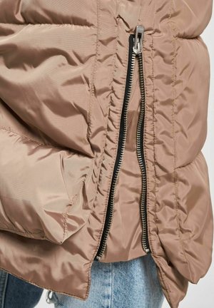 Částečně zipovaná béžová péřovka s viditelným stříbrným zipem přes světle modré džíny.