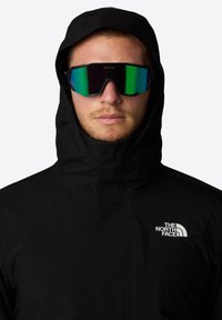 Zwarte waterdichte jas met hoge kraag en capuchon, voorzien van een wit logo op de borst en spiegellende groen-getinte zonnebril.