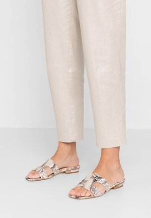 Pieds portant des sandales beige à enfiler avec motif peau de serpent et petits talons blocs, associées à un pantalon crème clair longueur cheville.