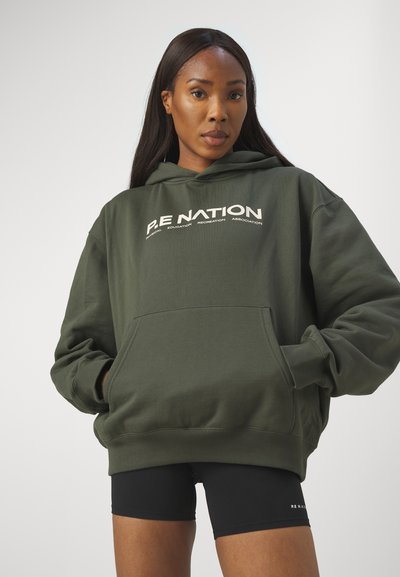 Felpa verde oliva con tasca frontale, caratterizzata dal logo bianco "P.E Nation". Tessuto morbido, vestibilità oversized, polsini e orlo a costine. Pantaloni corti neri visibili.