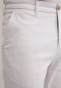 Pantaloni grigi chiari realizzati in misto cotone, con tasche laterali, zip e chiusura a bottone in legno. Texture liscia con un design aderente.