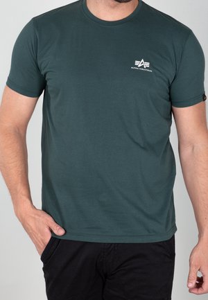 Ein Mann trägt ein dunkelgrünes Alpha Industries T-Shirt mit Logo auf der Brust und schwarze Hose, steht mit einer Hand in der Tasche.