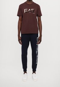 Ruskea puuvillainen t-paita, jossa on kontrastivärinen logo, yhdistettynä laivastonsinisiin collegehousuihin, joissa on valkoiset brändäykset sivulla. Valkoiset tennarit viimeistelevät asun.