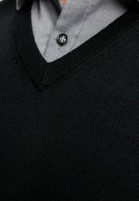 Schwarzer V-Ausschnitt-Pullover mit strukturiertem Strick, über einem hellgrauen karierten Hemd mit Knopfkragen getragen.