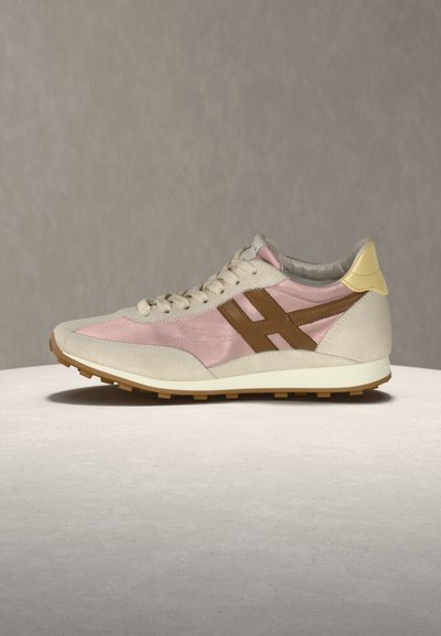 Χαμηλό sneaker με μπεζ σουέντ, ροζ υφασμάτινα πάνελ, καφέ λογότυπο στο πλάι, κρεμ σόλα και κίτρινη ταμπελίτσα στη φτέρνα πάνω σε ανοιχτή επιφάνεια με γκρι φόντο.